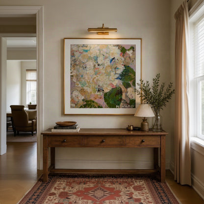 A large framed abstract floral painting hangs on a beige wall above a wooden console table in a styled interior. The artwork features soft cream, blush, green, and blue tones with a gold frame and a brass wall sconce above it.